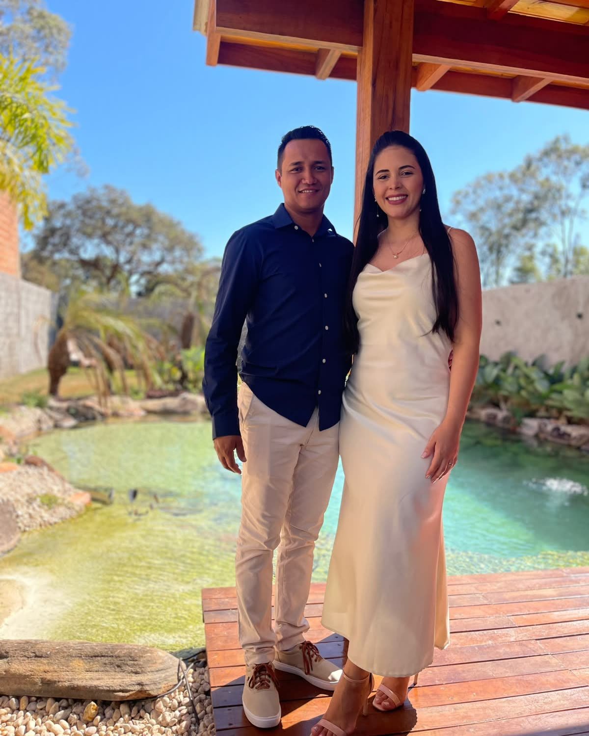 Casal recém-casado no deck de madeira sob o pergolado, com o lago ao fundo